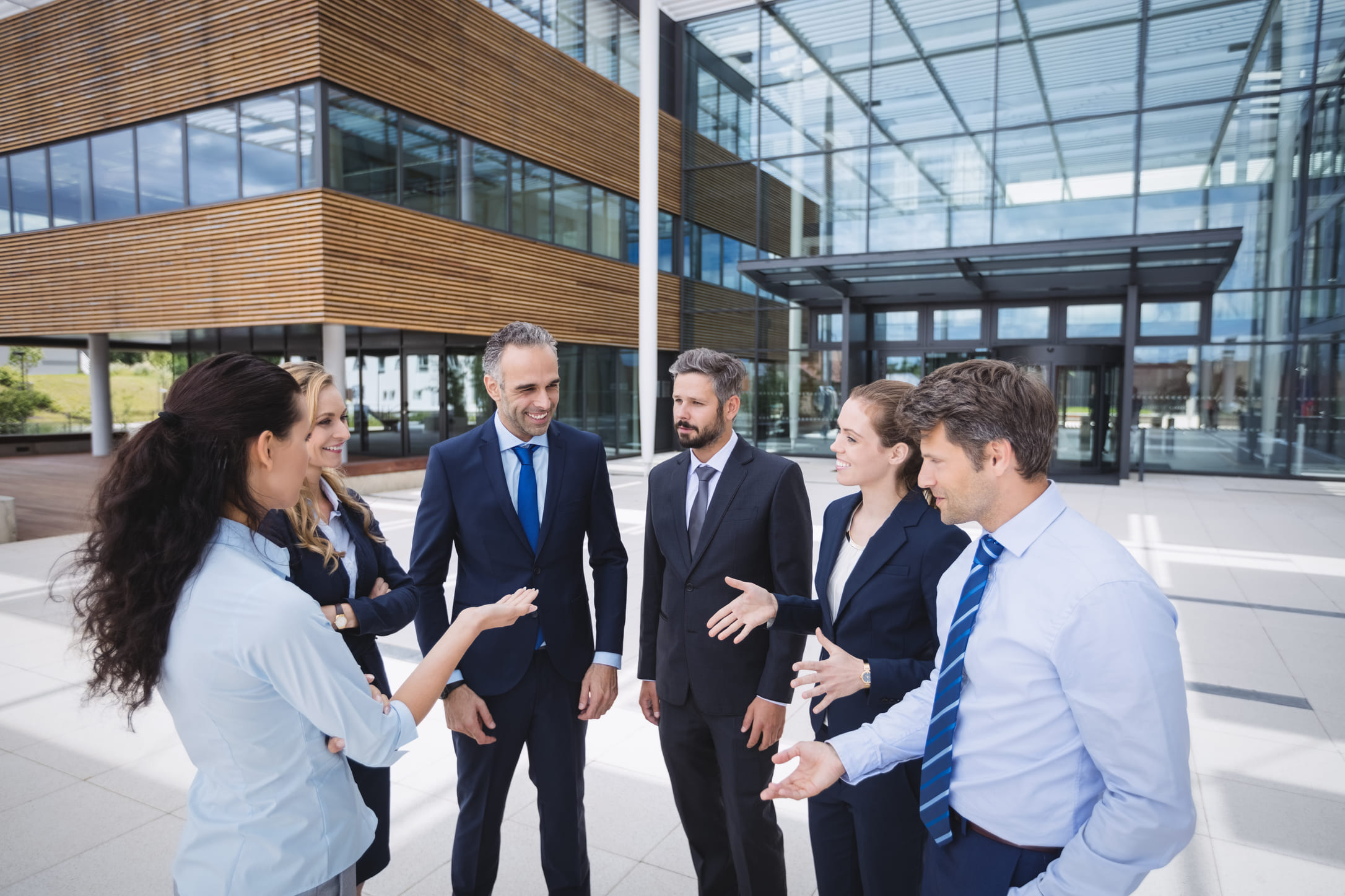 Business professionals in suits and smart-casual attire converse outdoors near a modern glass office building, conveying collaboration and confidence.