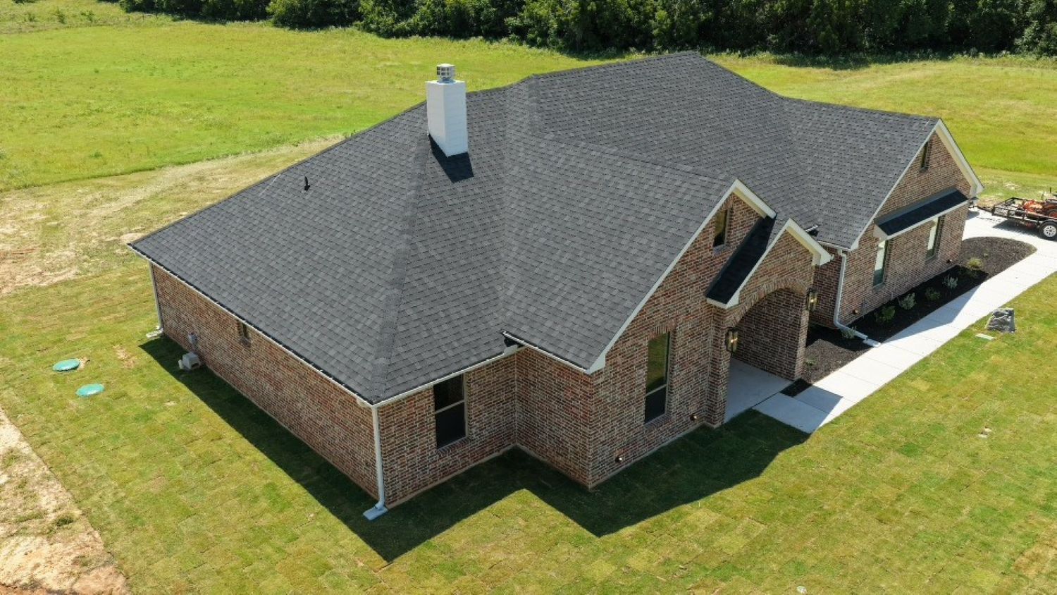 Aerial view of a large brick suburban home with a dark shingle roof, chimney, driveway, and expansive green lawn in bright daylight.