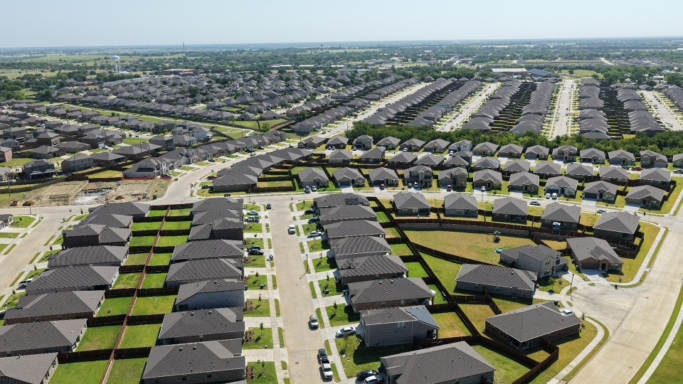 Aerial view of a sprawling suburban neighborhood with rows of modern homes, curving streets, green lawns, and distant farmland.