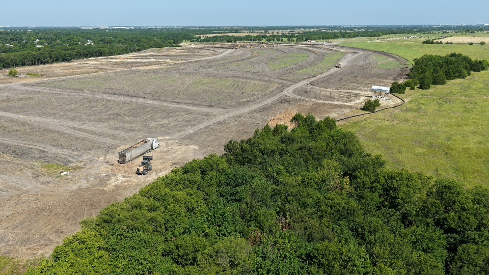 Aerial view of a vast cleared construction site bordered by dense trees and green fields, with a dump truck and excavator on the dirt.