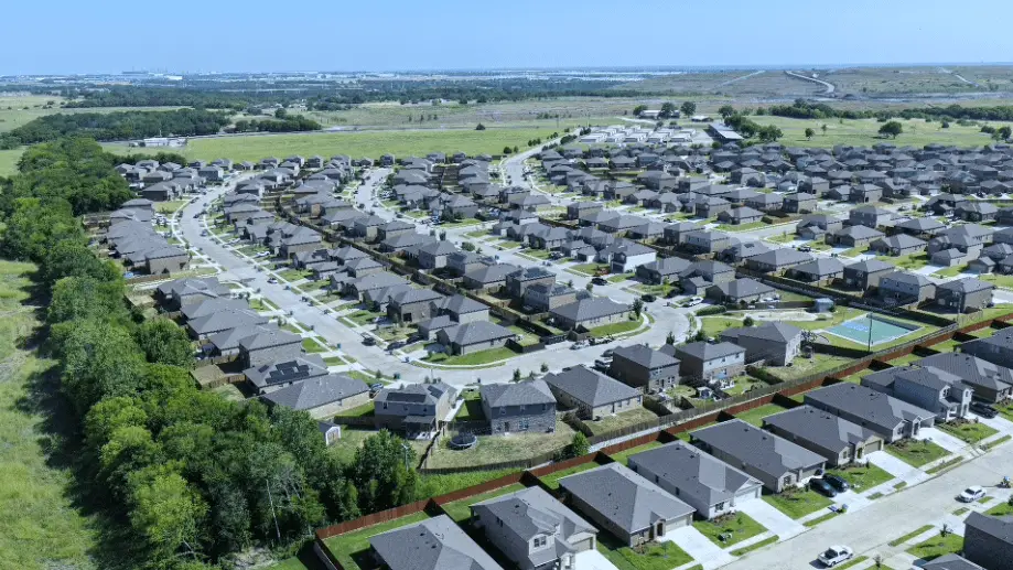 Aerial view of a sprawling suburban housing development with rows of gray-roofed homes, curving streets, green lawns, and distant fields.