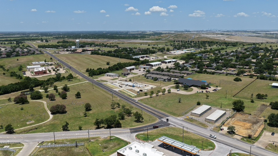 Aerial view of a rural town with roads, homes, fields, farm buildings, and an industrial complex under a bright, peaceful blue sky.