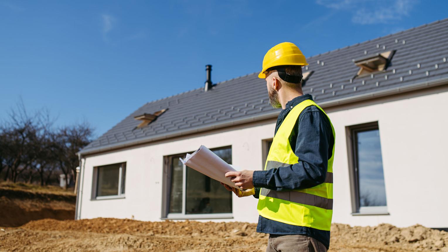 Construction worker in a yellow hard hat and safety vest reviews blueprints outside a newly built house under a clear blue sky.